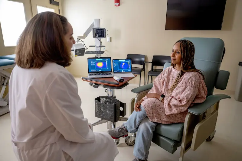 A healthcare professional in a white coat speaks with a patient in a pink hospital gown, while two laptops display medical imaging on a rolling cart.