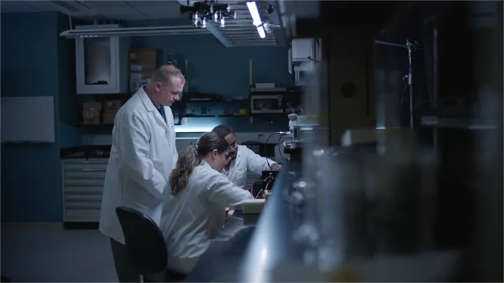 Three scientists in white lab coats work in a dimly lit laboratory, with one man observing two others who are focused on benchtop equipment.