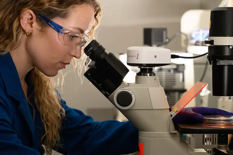 A female scientist in a blue lab coat and safety glasses looks into a microscope, with a petri dish under the lens.