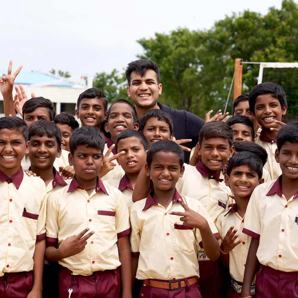 A smiling man in a green Notre Dame sweatshirt stands near a group of young children in blue and yellow uniforms outdoors.