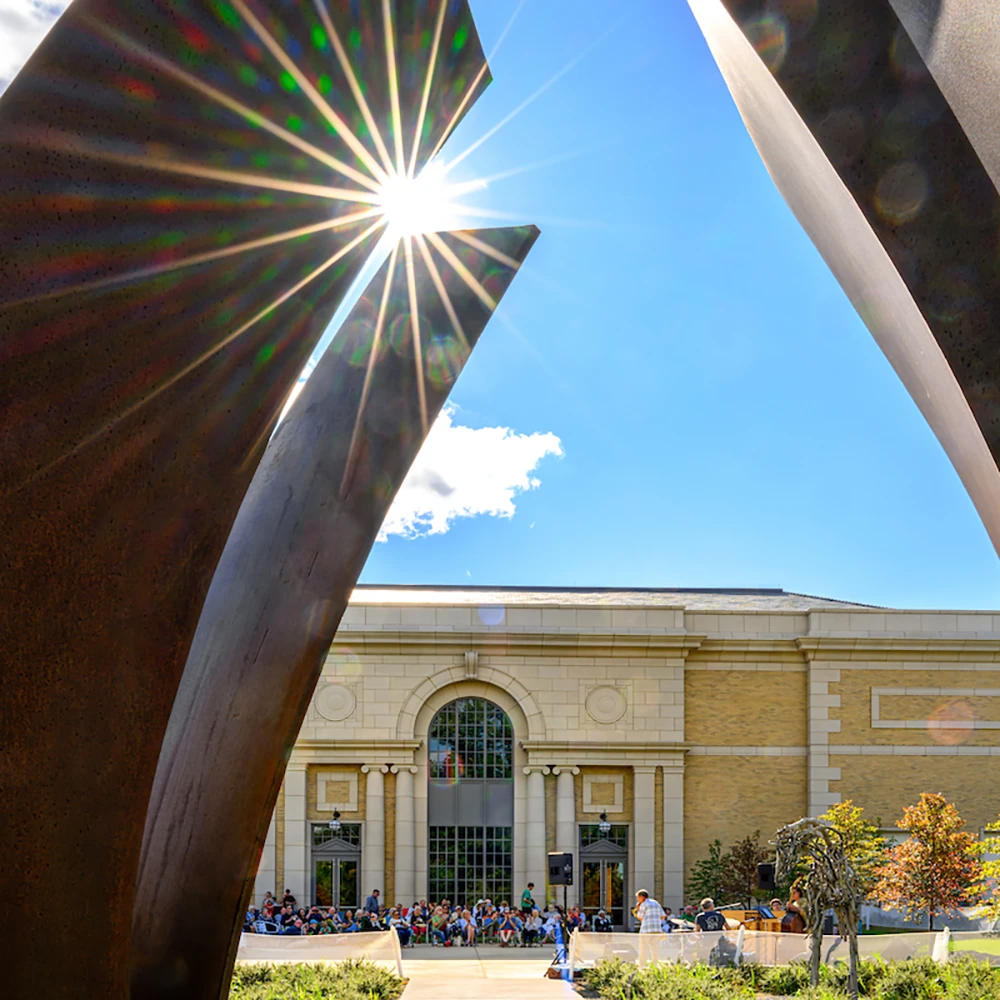 A low-angle view of the Raclin-Murphy Museum of Art, framed by a rust-colored sculpture, with a crowd gathered outside on a sunny day.