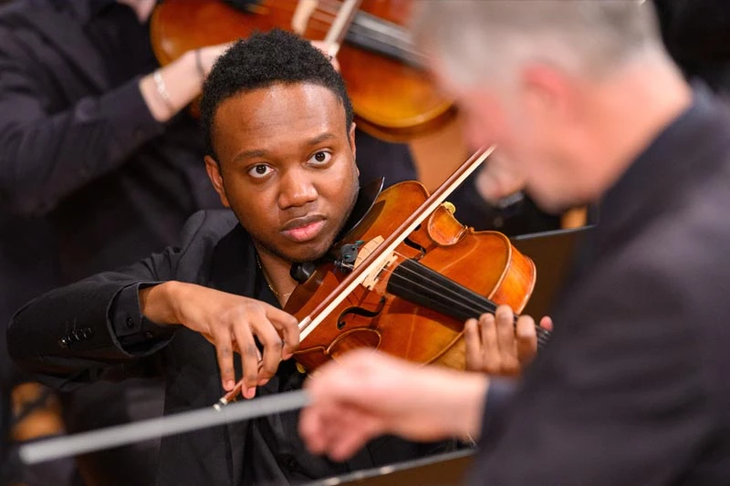A Black male violinist in a black suit looks forward while playing his instrument in an orchestra.