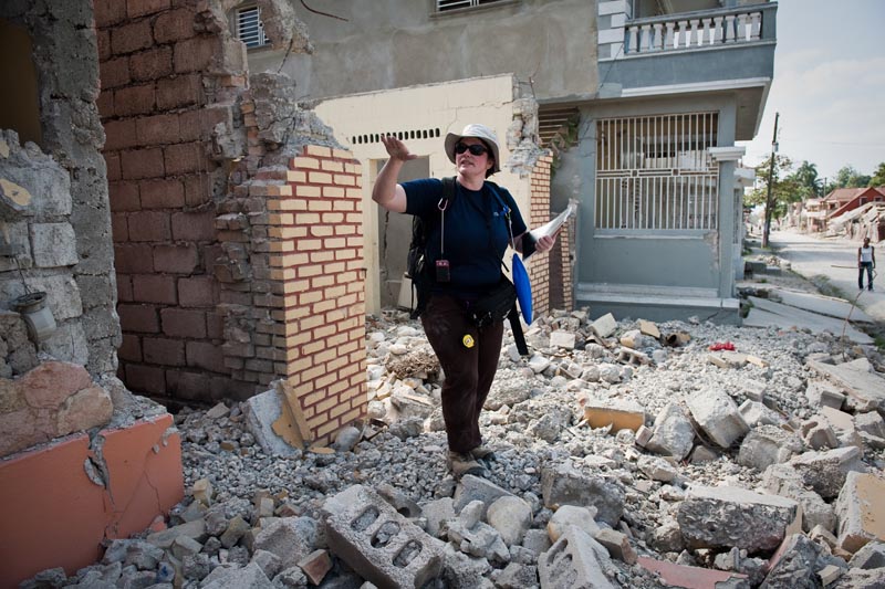 A woman in a hat and backpack stands in a rubble-filled street, gesturing as she surveys the damage.