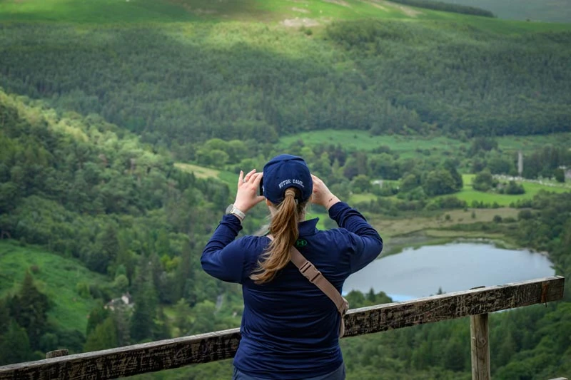 A person wearing a Notre Dame hat stands at a wooden railing, looking out over a green valley with a lake.