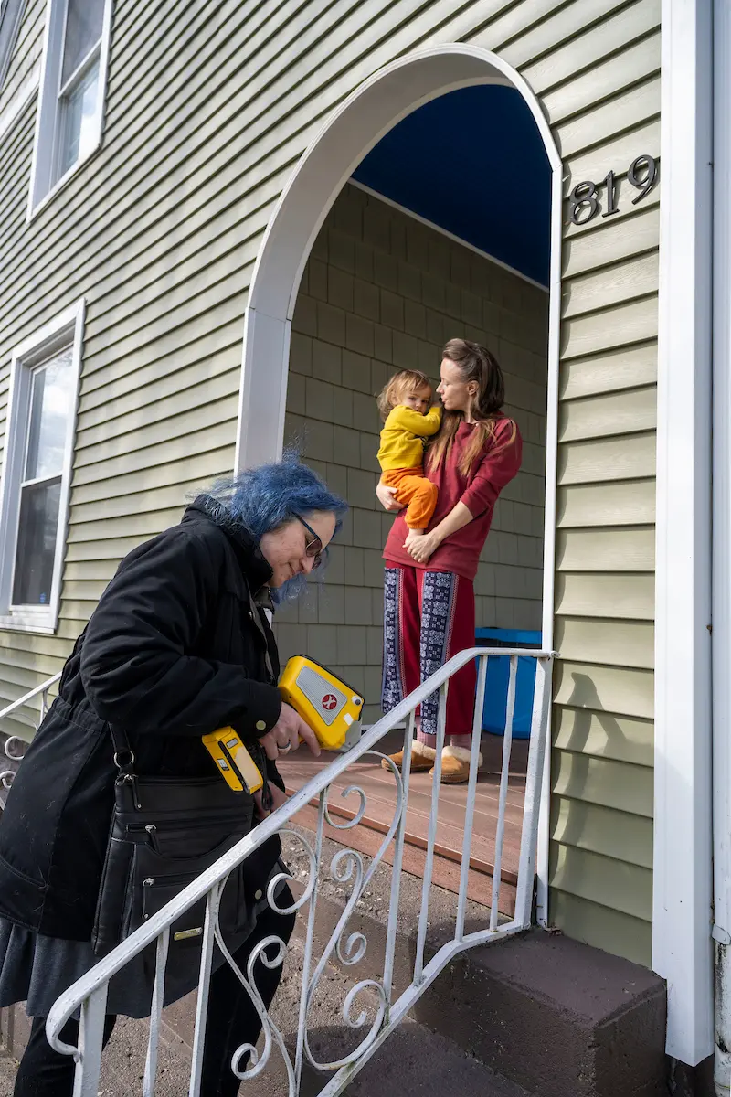 A woman with blue hair, wearing a black coat, looks down at a device while standing on steps, with a woman holding a child in a doorway behind her.