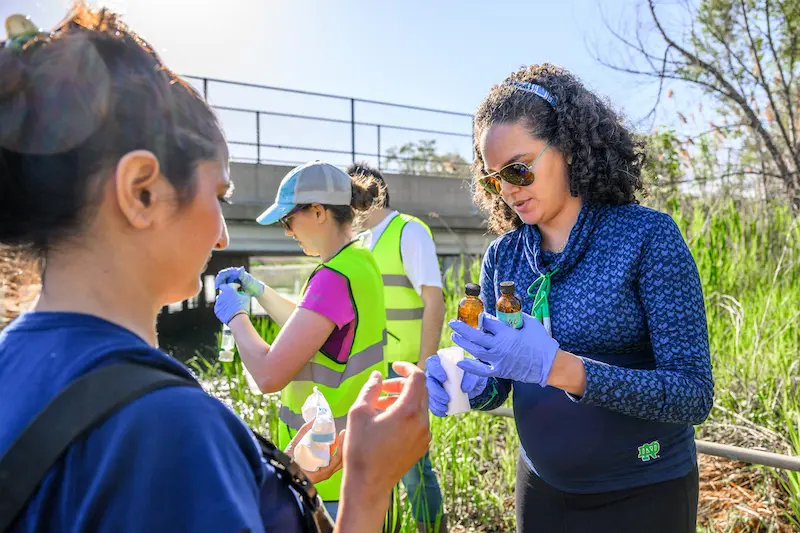 A female researcher in sunglasses and blue gloves examines liquid samples with another woman, while a third person in a safety vest is in the background near a bridge.