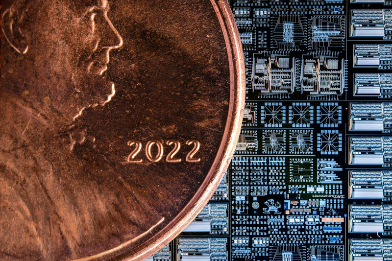 The edge of a 2022 copper penny is magnified, juxtaposed against the complex grid pattern of a computer microchip.