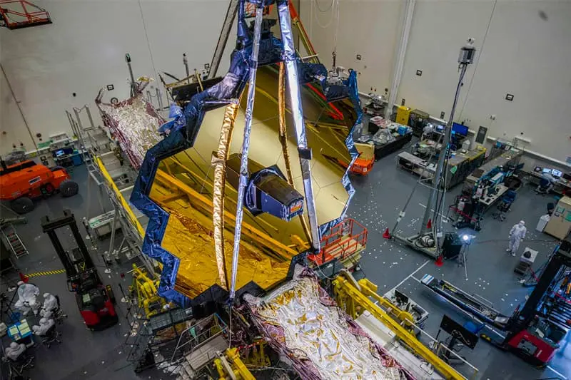 High-angle view of the James Webb Space Telescope's gold-colored hexagonal mirrors being assembled in a large clean room.  Support equipment and personnel in clean suits are visible below.