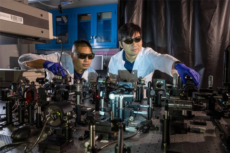 Two researchers in white lab coats and protective eyewear work with a complex optical setup on a metal table.  They wear purple gloves and are adjusting components of the experiment.